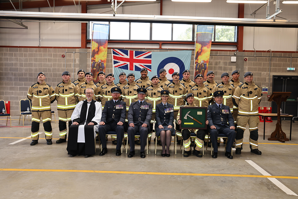 A group photograph inside a large hanger features graduating firefighters posed in front of a Union Jack flag and a Royal Air Force flag. In the front row, four training staff in formal dark blue uniforms and chaplain are seated. Standing behind them are two rows of graduating firefighters wearing tan and yellow firefighting gear with berets.